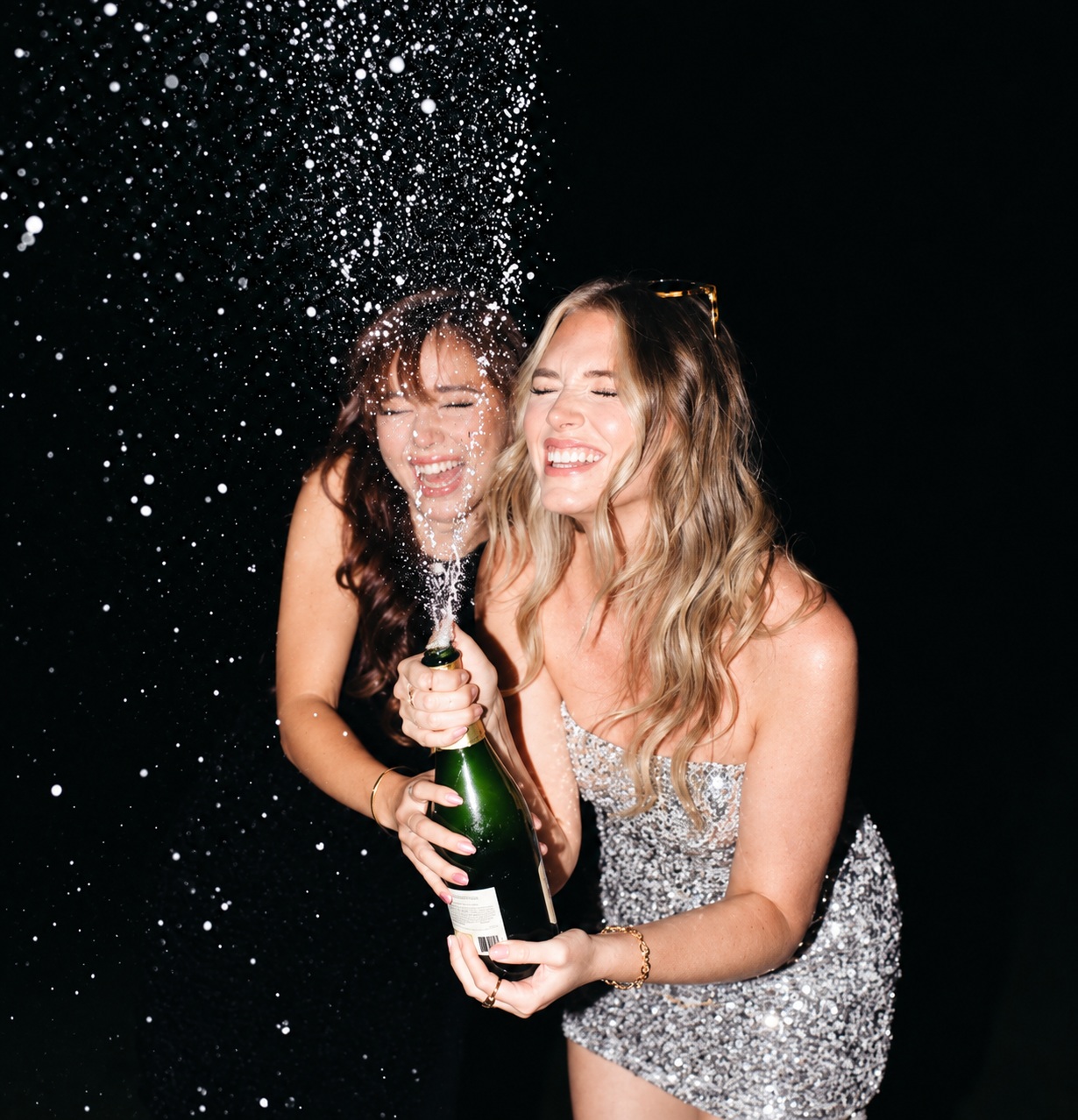Two friends laughing as a champagne bottle sprays against a black background