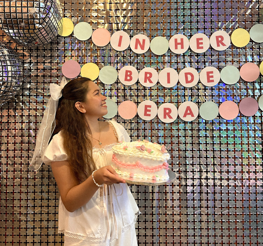 A bride to be in a bow veil holding a heart cake in front of an In Her Bride Era sign on a disco wall