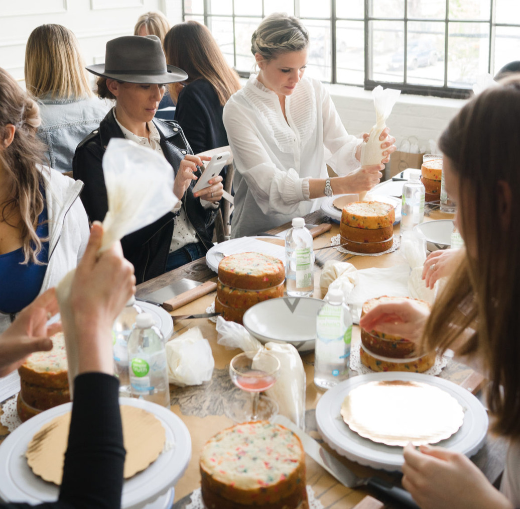 Friends at a long table with piping bags and funfetti layer cakes on turntables