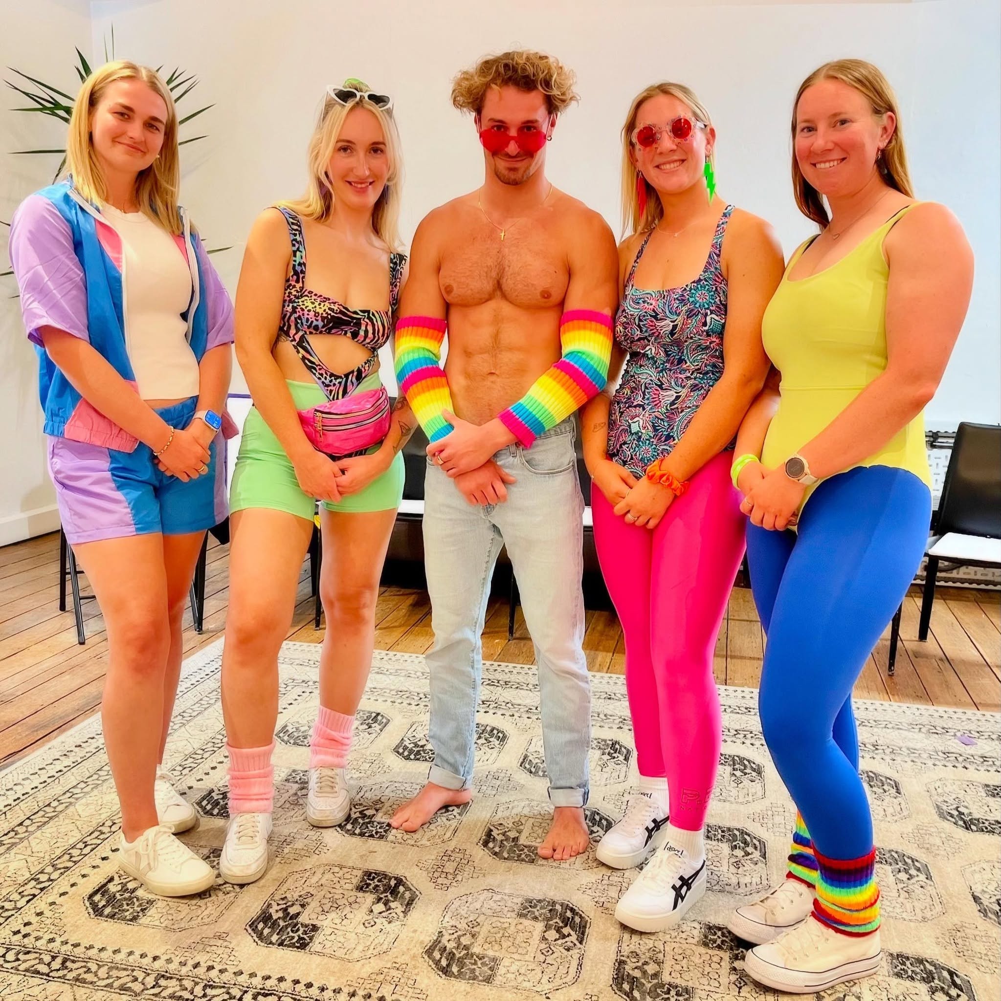 Four hens in eighties leotards and leg warmers posing with a shirtless man in jeans