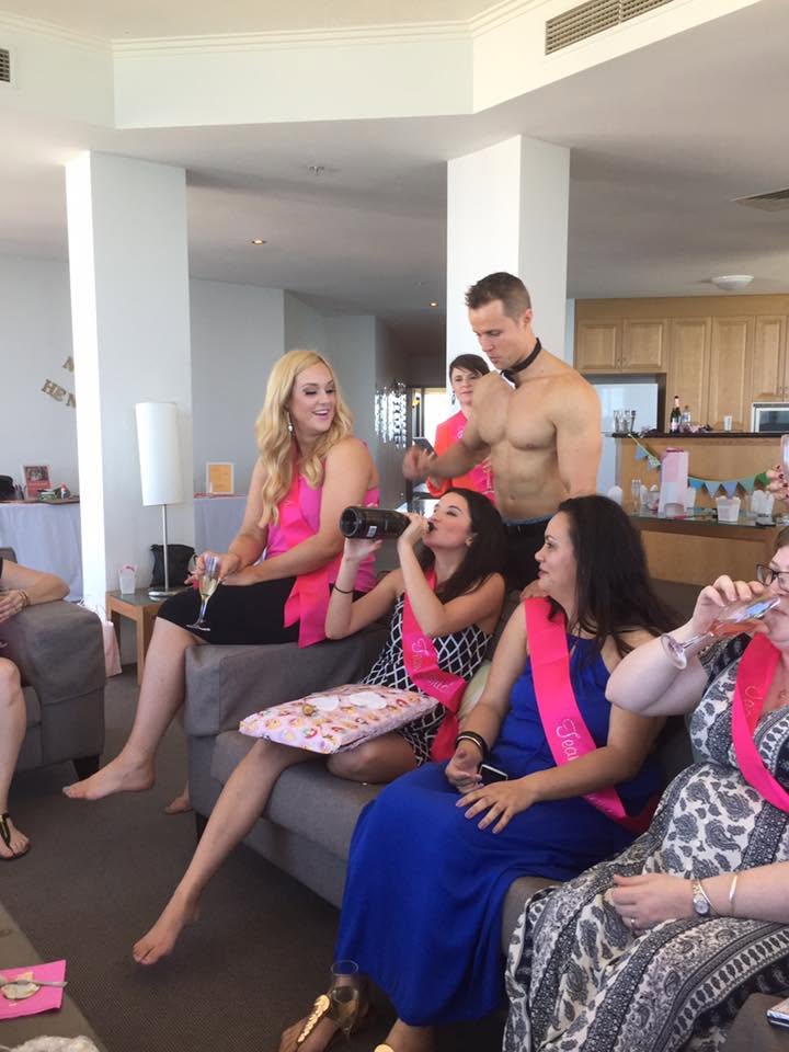 Hens in pink sashes with glasses of bubbly on a couch while our topless waiter in a bow tie looks after them