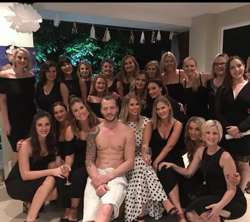 A big hens group in black dresses around the model, who sits wrapped in a white towel
