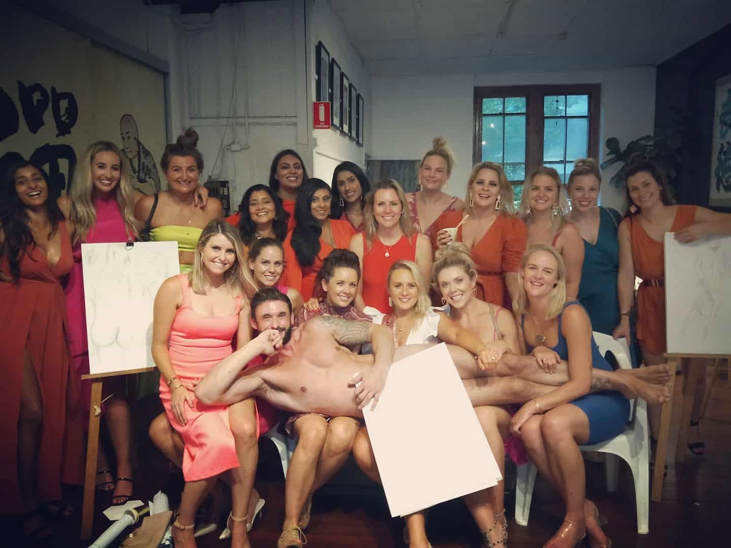 A big hens crew in bright dresses laughing in a drawing studio, the life model lying across their laps behind a sheet of paper, easels with their drawings either side