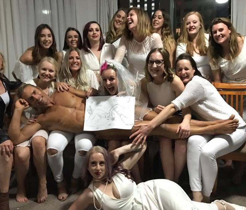 A hens group all in white, the bride in a veil, with the model across their laps holding a sketch