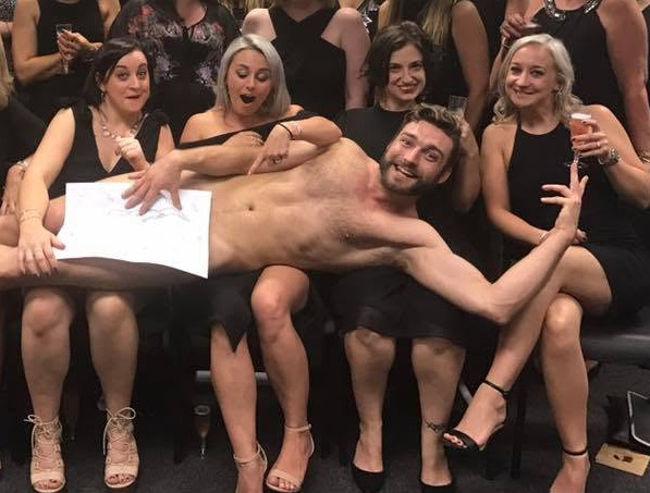 Hens in black dresses with the model lying across their laps, a drawing held over him