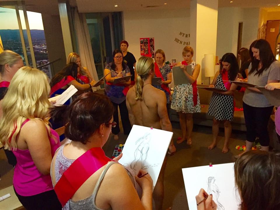 Hens in pink sashes standing in a circle with sketch pads, drawing the life drawing model in the middle of the room