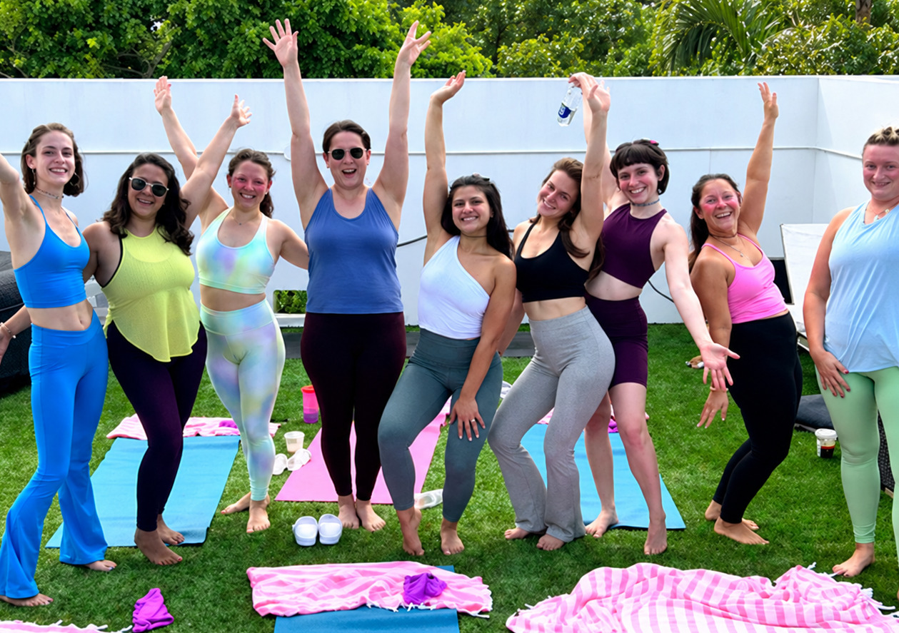 Nine friends in activewear with their arms raised beside yoga mats on a garden lawn