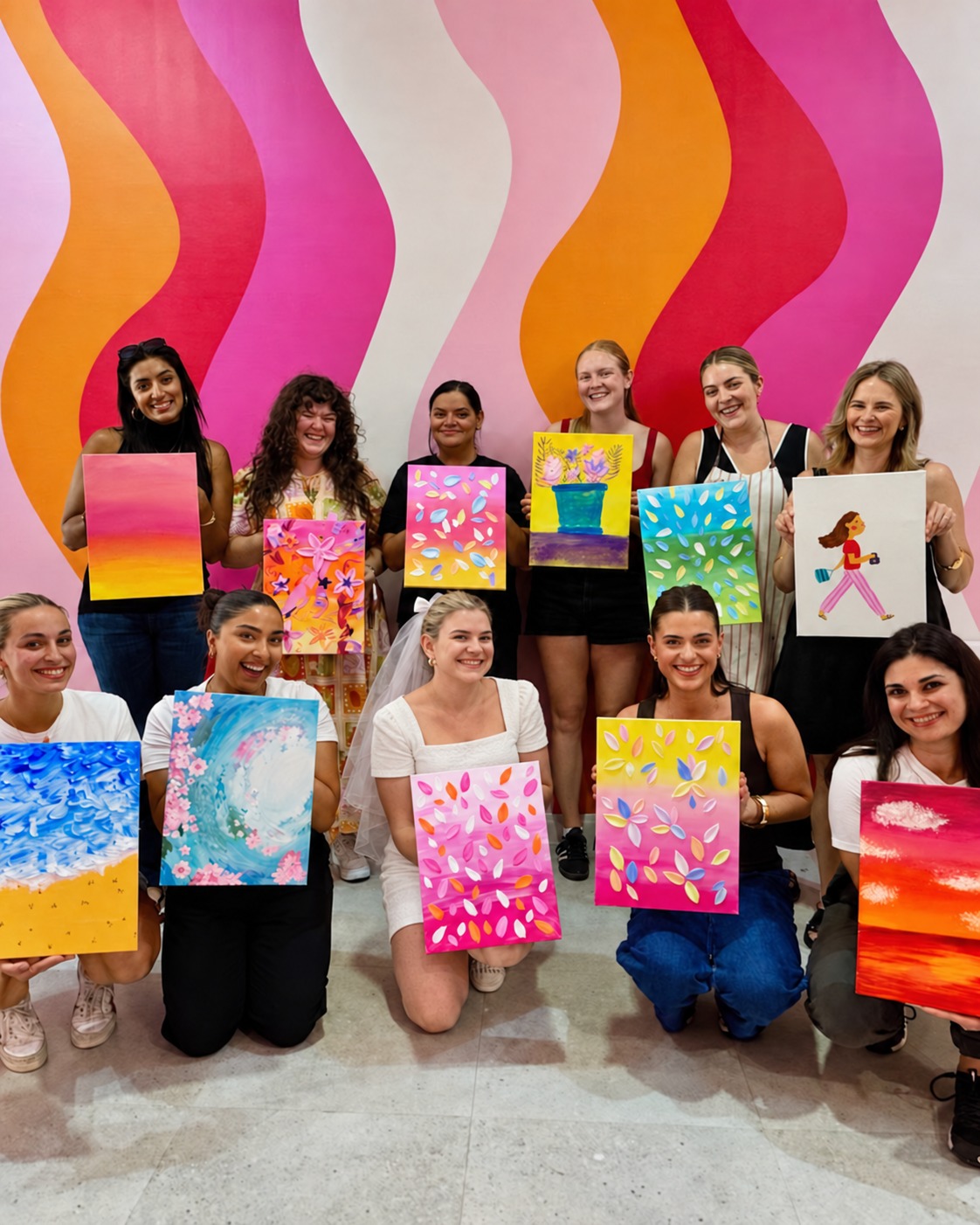A hens crew holding up their finished canvases in front of a wavy pink and orange mural, the bride in white and a veil kneeling in the centre