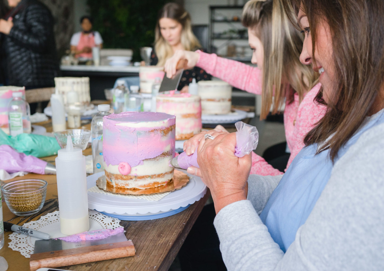 A woman piping pink buttercream onto a naked cake while her friends decorate cakes behind her