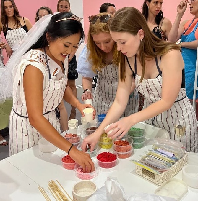 The bride in an apron and sash scoops coloured pieces with two friends at a table of colour pots