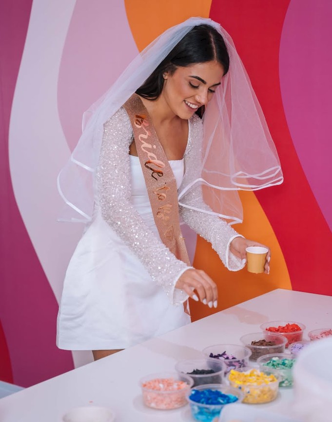 The bride in beaded sleeves, a veil and a sash smiles over pots of coloured pieces, a cup in hand