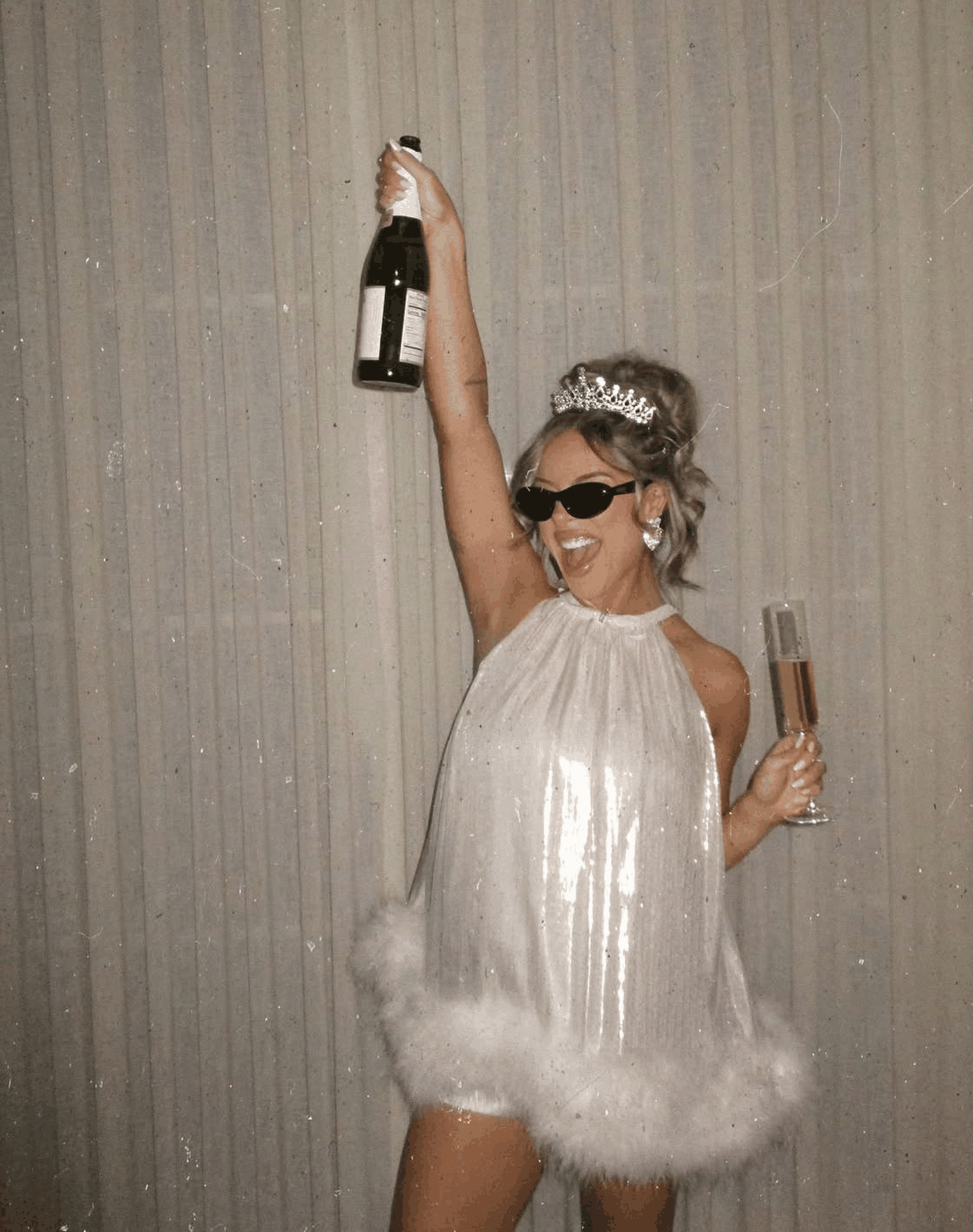 The bride in a white feather dress and tiara holding a bottle of bubbles over her head