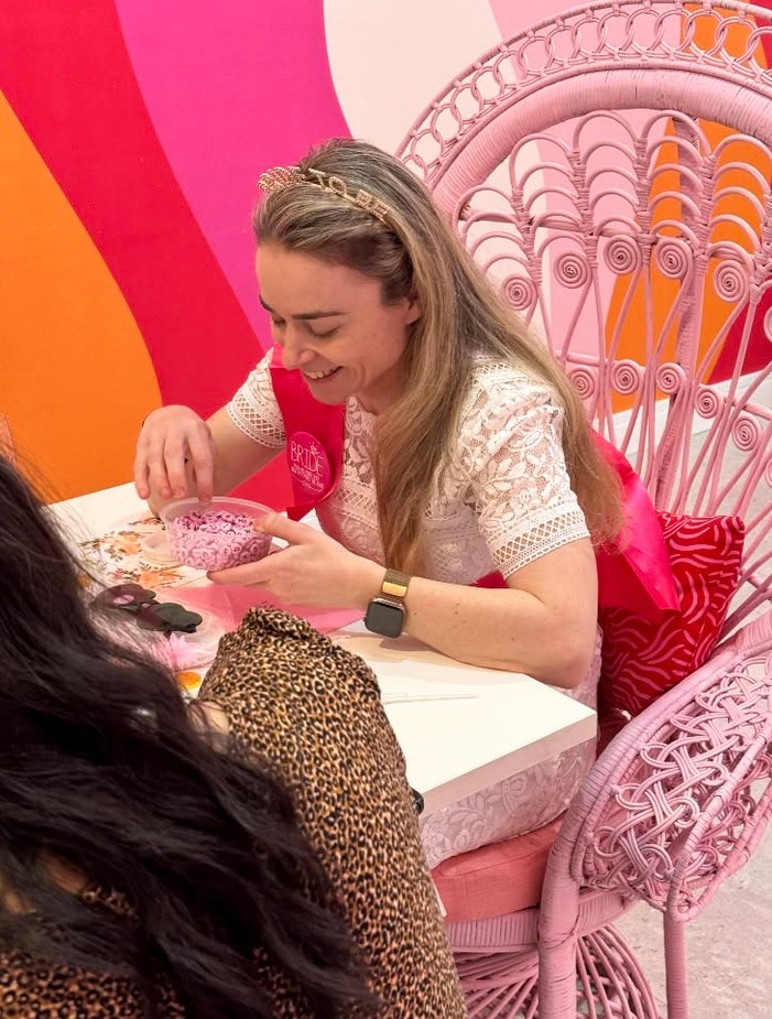 The bride in a sash and tiara laughs as she scoops tiny pink beads from a bowl, sitting in a pink peacock chair