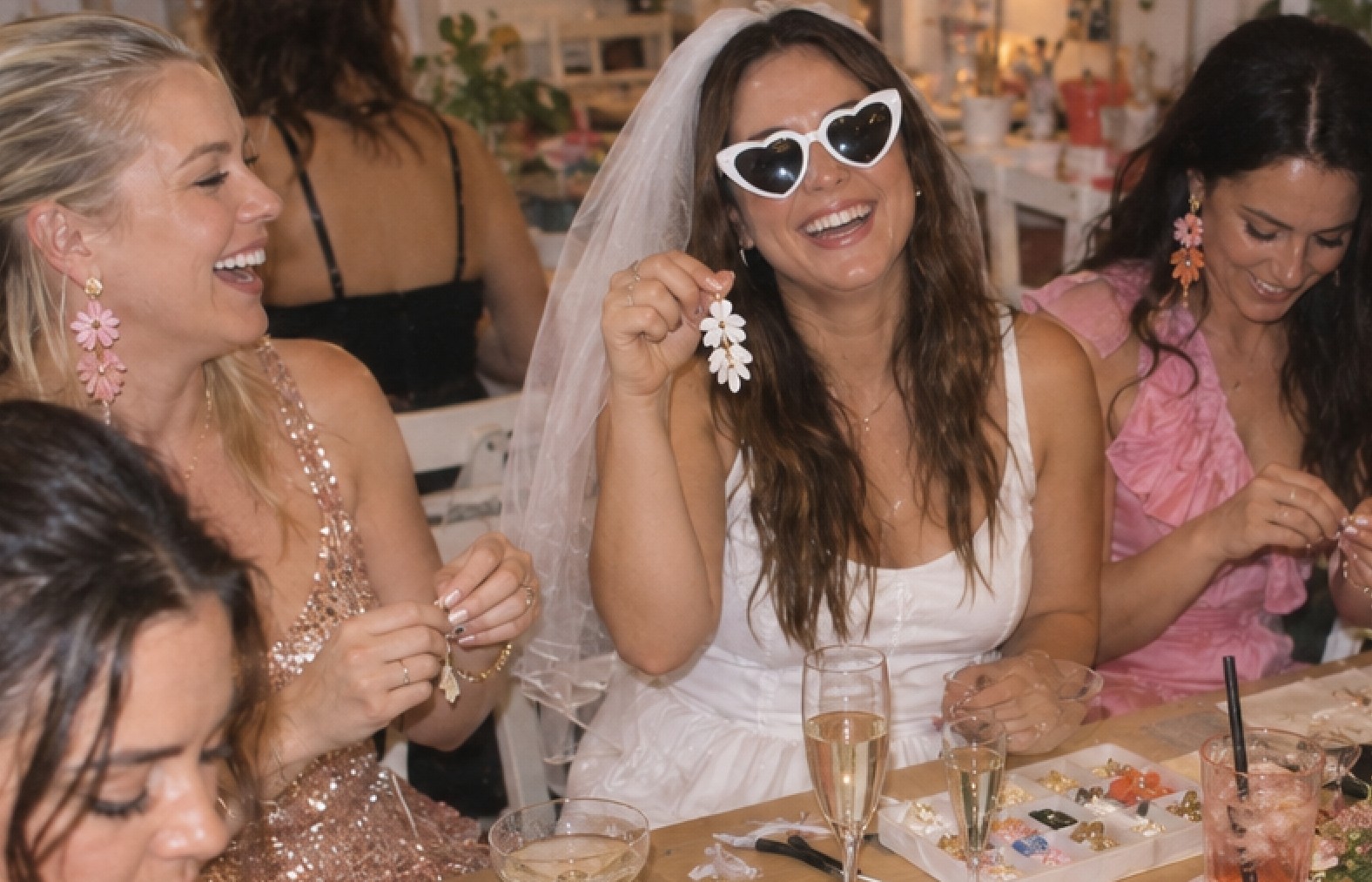 The bride in a veil and white heart sunglasses laughs as she holds up the white flower earring she made, her friends wearing their pink flower earrings