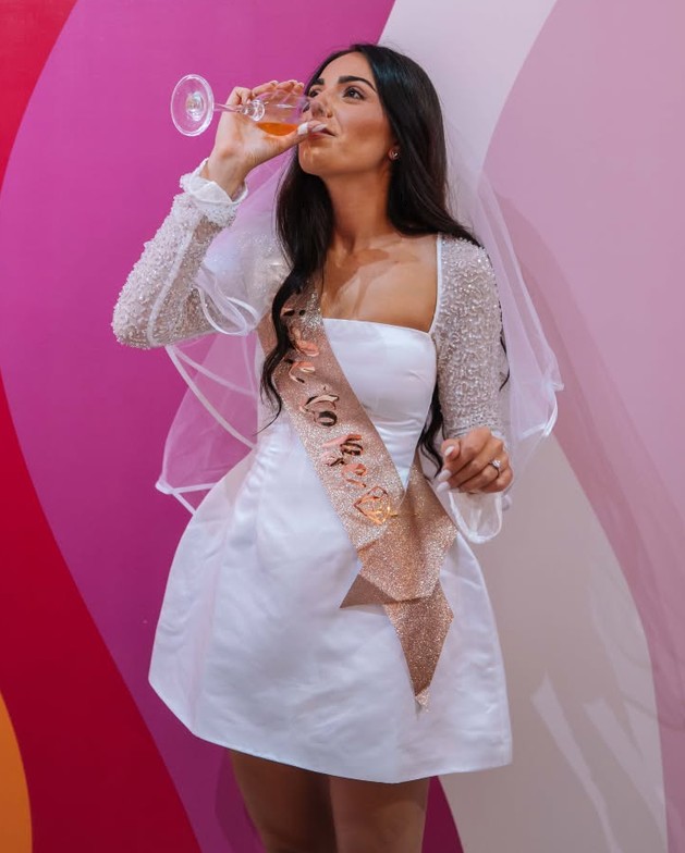 The bride in a white dress with beaded sleeves, a veil and a sash sips a glass of bubbly
