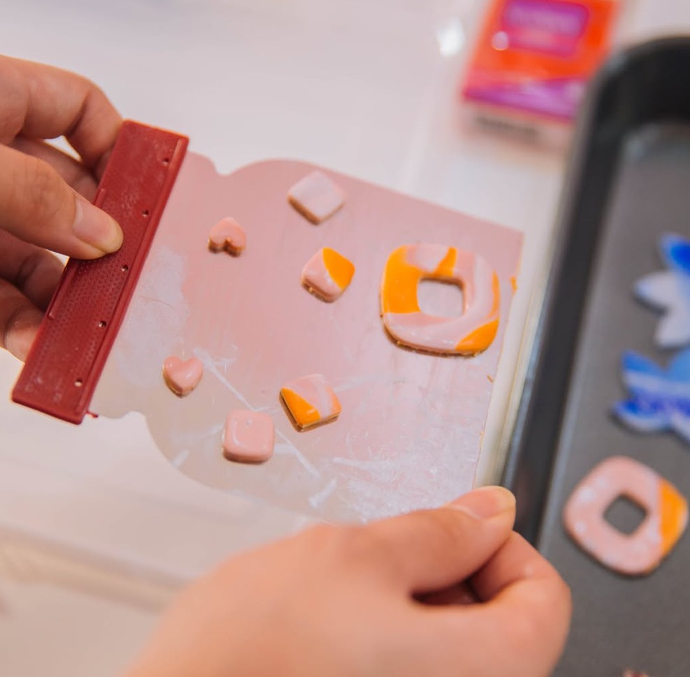 Hands lift pink and orange clay earring pieces on a tile beside a tray of blue clay flowers