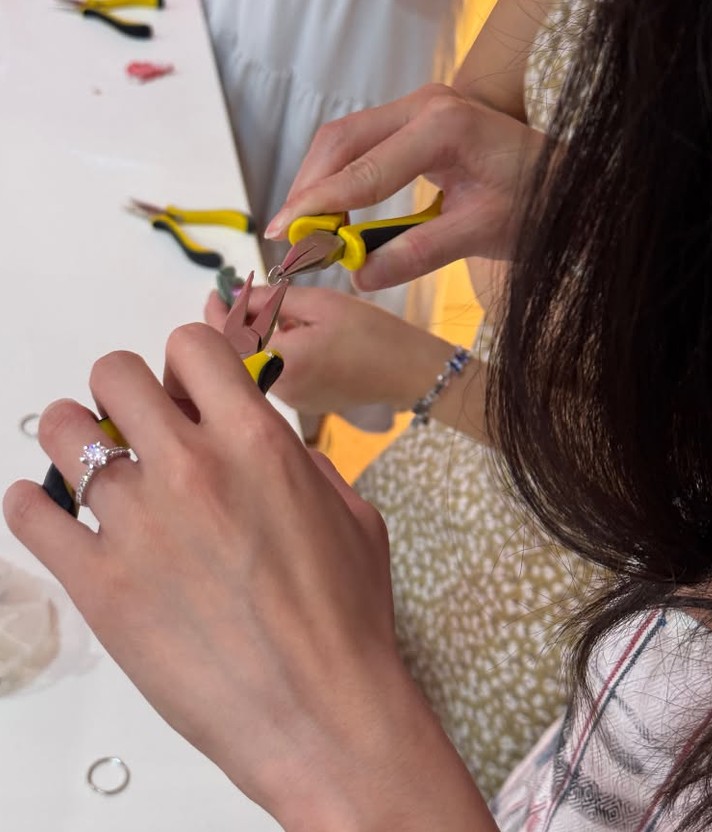 A close-up of a hand wearing an engagement ring holds pliers to open a silver jump ring while a friend works beside her