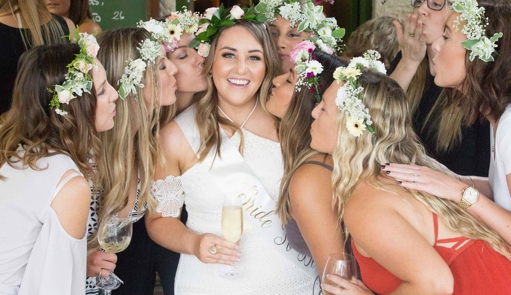 The bride in her sash laughing as her friends in fresh flower crowns kiss her cheeks