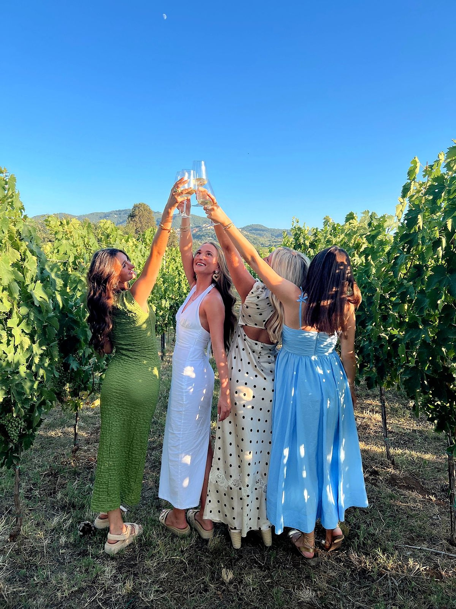 Four friends in summer dresses raising their glasses together in a vineyard under a blue sky