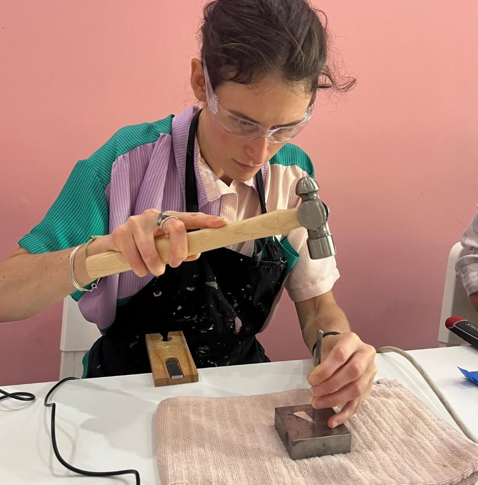 A guest in safety glasses brings a hammer down on a metal stamp at a steel block
