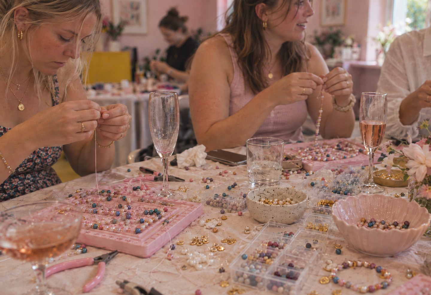 Two friends thread pearl and coloured glass beads at pink bead boards, with glasses of rose bubbly and bowls of gold spacers