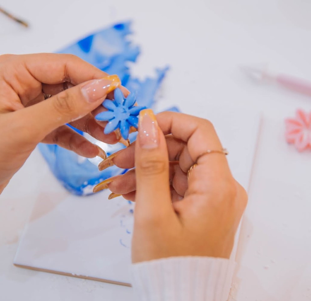 Hands with long nails shape a blue clay flower earring over a tile
