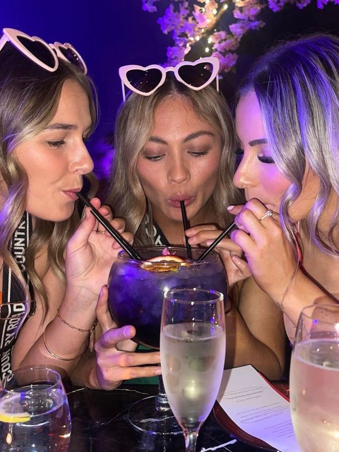 Three friends in heart sunglasses sharing a purple fishbowl cocktail through straws