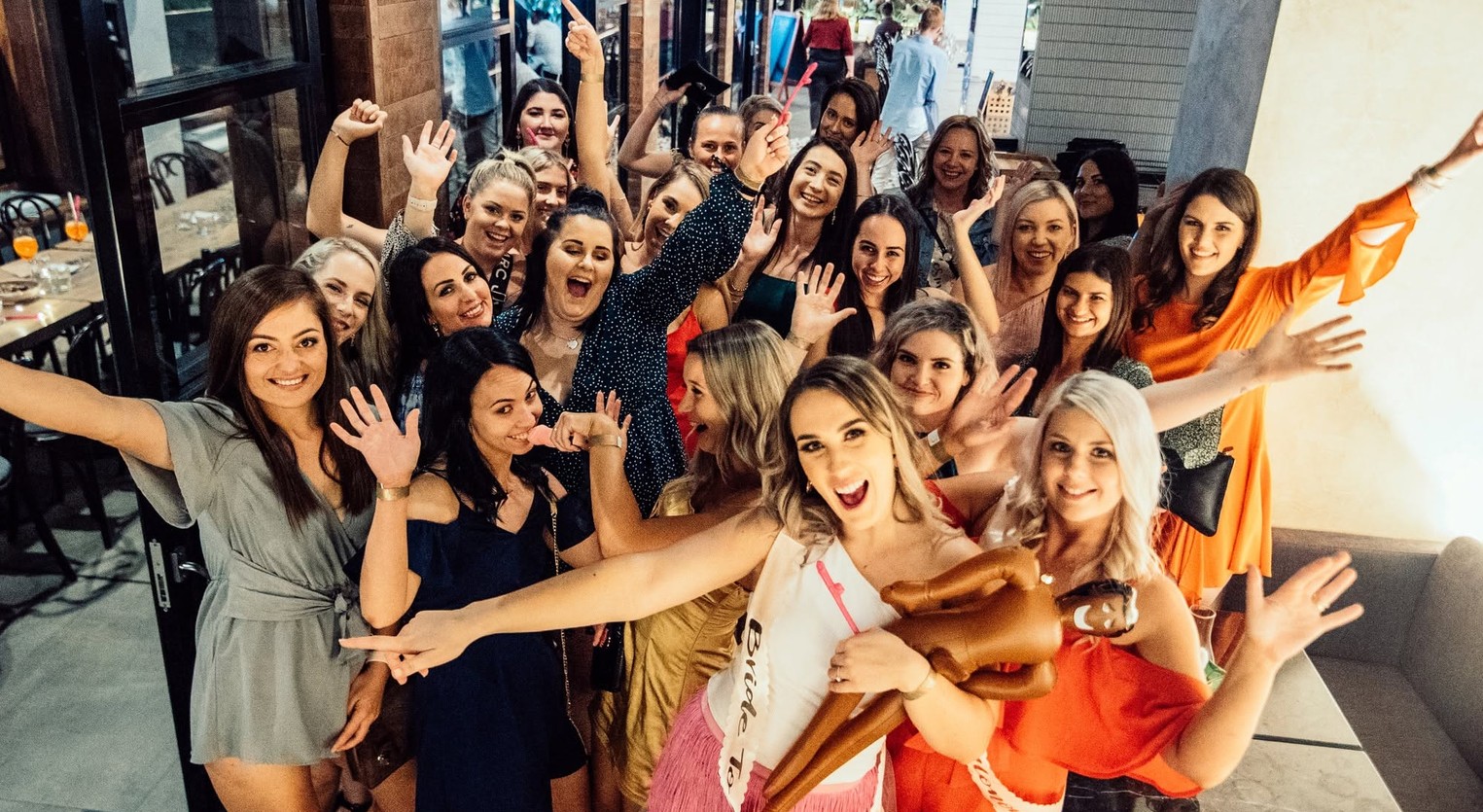 A big hens crew in a restaurant with every hand in the air, the bride in front in her sash hugging an inflatable man