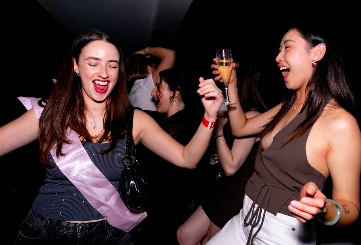 The bride in her sash laughing mid-dance at the club with a friend holding a glass of bubbles