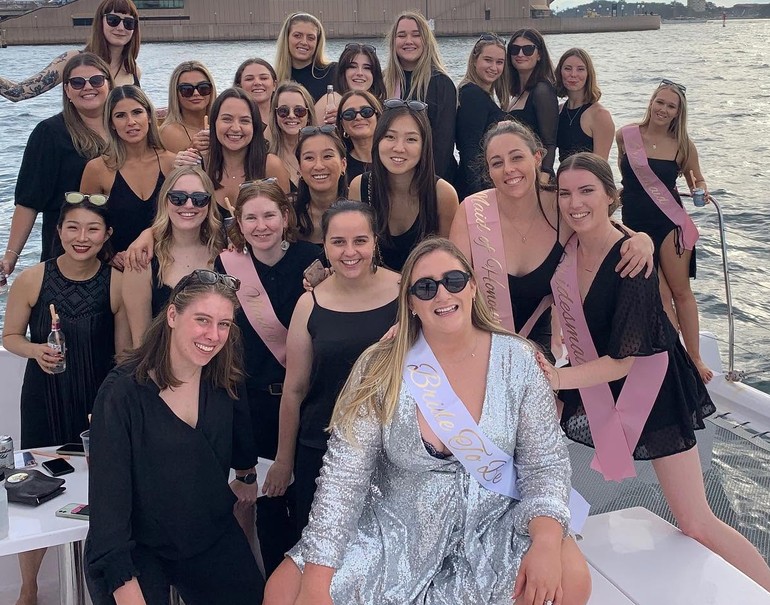 A big hens crew in black and pink sashes on the deck of a catamaran on the harbour, the bride front and centre in silver sequins