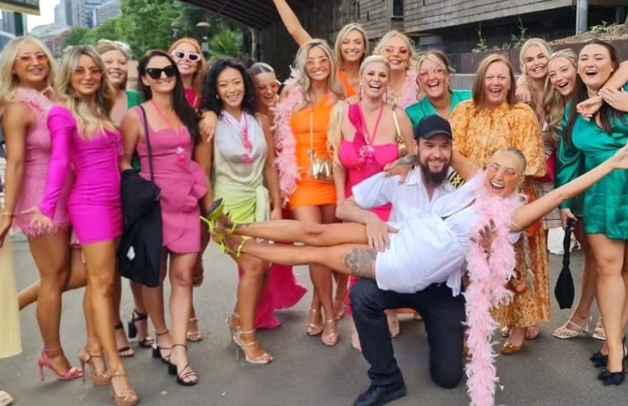 A big hens crew in bright dresses and pink boas laughing together on the wharf, one hen held up for the photo