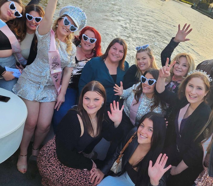 The bride in silver sequins and a sparkly captain hat with her crew in heart sunglasses, everyone waving from the deck of a river cruise