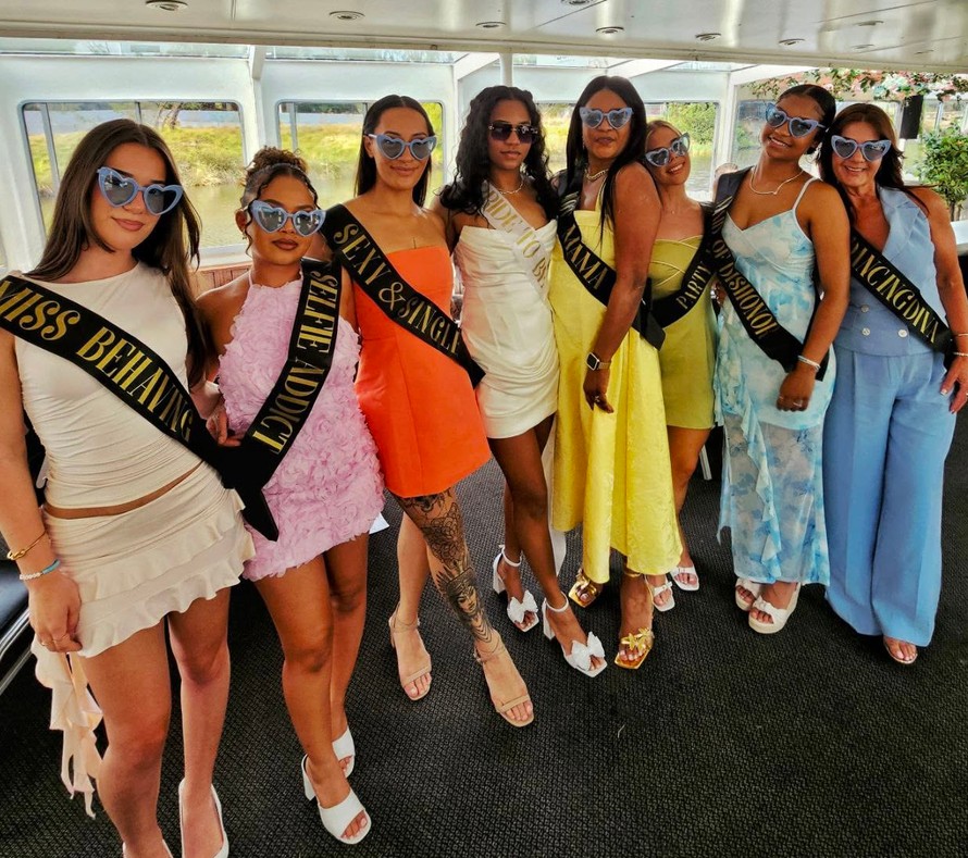 A hens crew in black sashes and blue heart sunglasses lined up inside a river boat, the bride in white in the middle