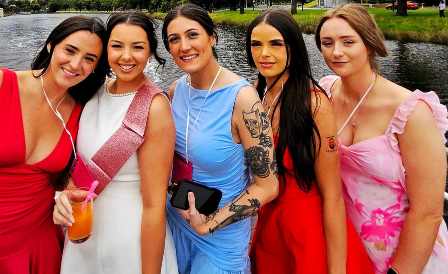 The bride in white with a pink sash and four friends in bright dresses by the river