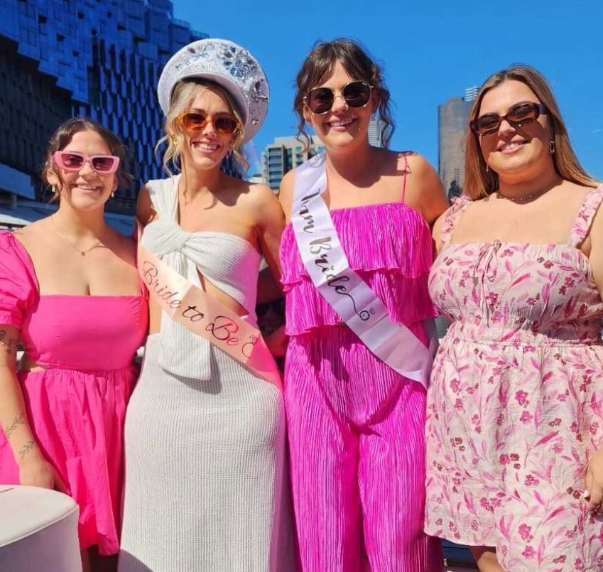 Four friends in pink on a sunny city deck, the bride in her sash in the middle