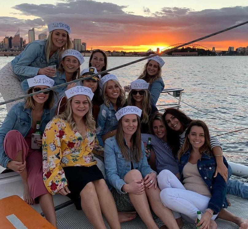 A hens crew in I do crew sailor hats on a catamaran net at sunset, the harbour bridge and a pink sky behind them