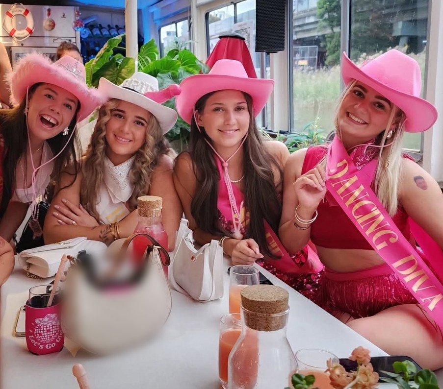 Four friends in pink cowgirl hats and sashes laughing at a table on board, pink cocktails in front of them