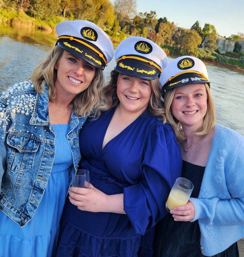 Three friends in captain hats smiling together on deck in the late afternoon light, the river behind them