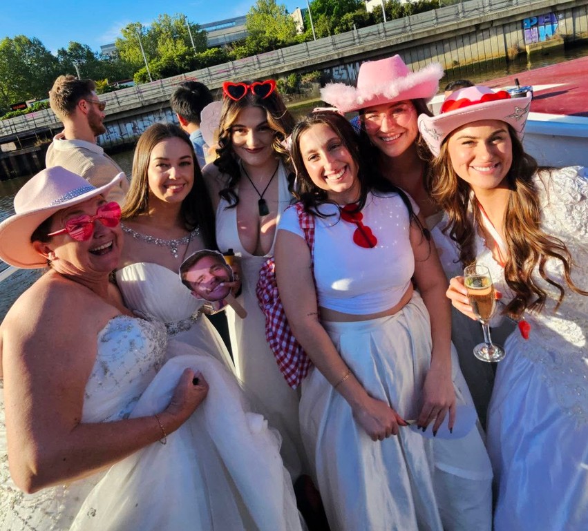 A crew in white dresses and pink cowgirl hats on the river, one friend laughing in pink heart sunglasses