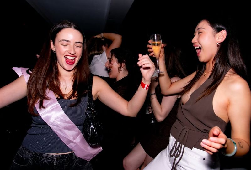 Two hens dancing and laughing on a club dance floor, one in a sash, one holding a glass of bubbles