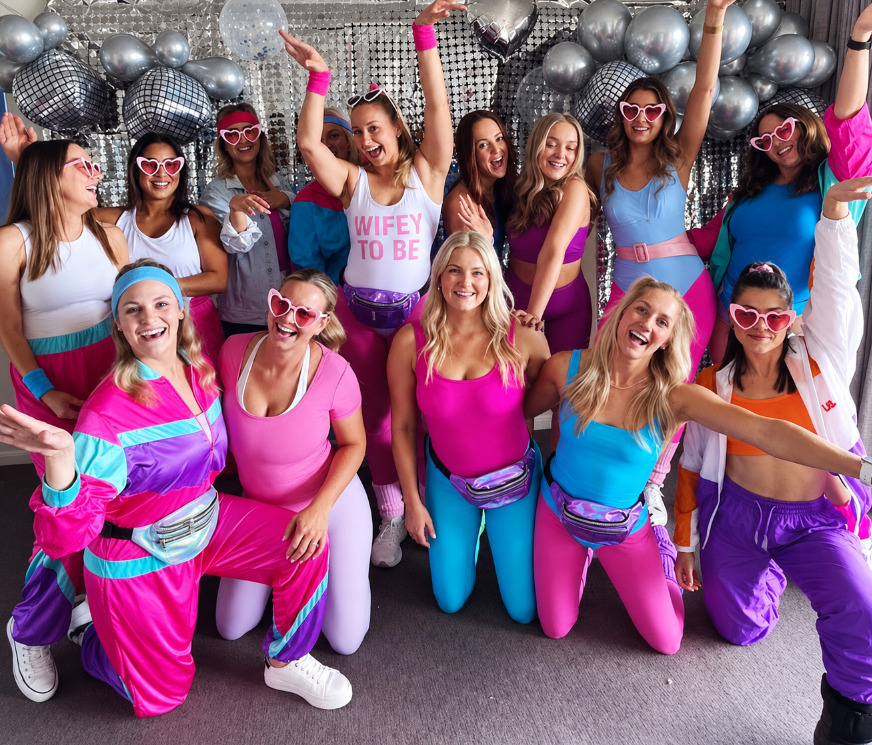 A hens group in 80s-style shell suits and heart sunglasses, the bride in a Wifey To Be top, with silver balloons