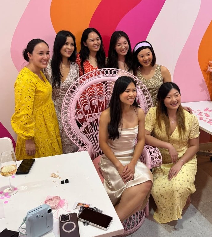 Seven friends gather round a girl in satin on a pink peacock chair