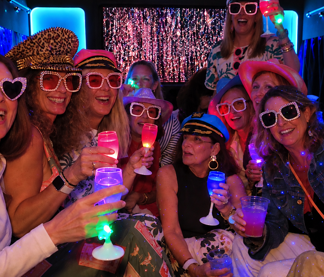 A big hens group in sparkly sunglasses and captain hats raising light-up cups on a party bus