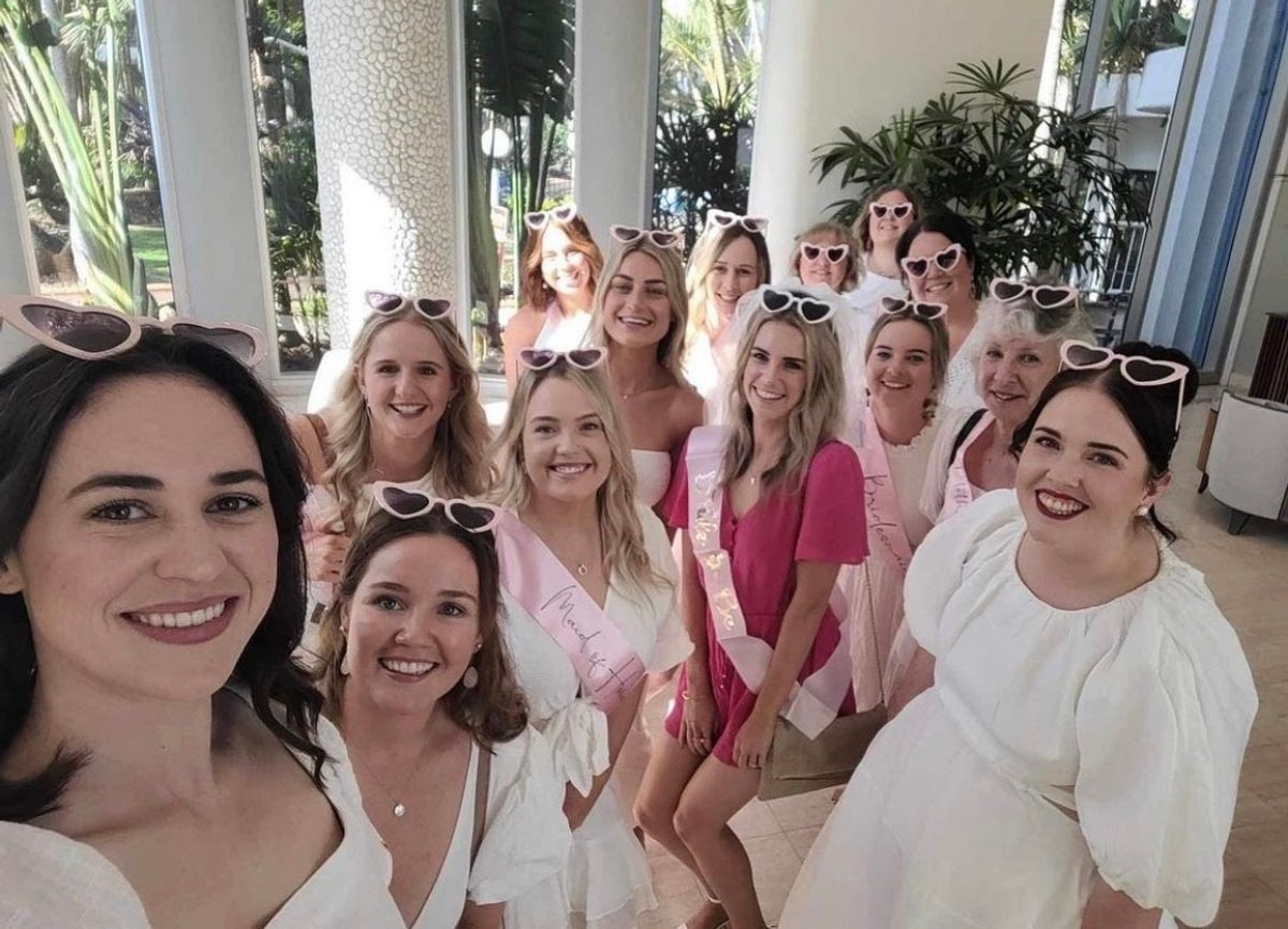 A hens group in heart-shaped sunglasses and summer dresses squeezing into a selfie at a bright daytime venue