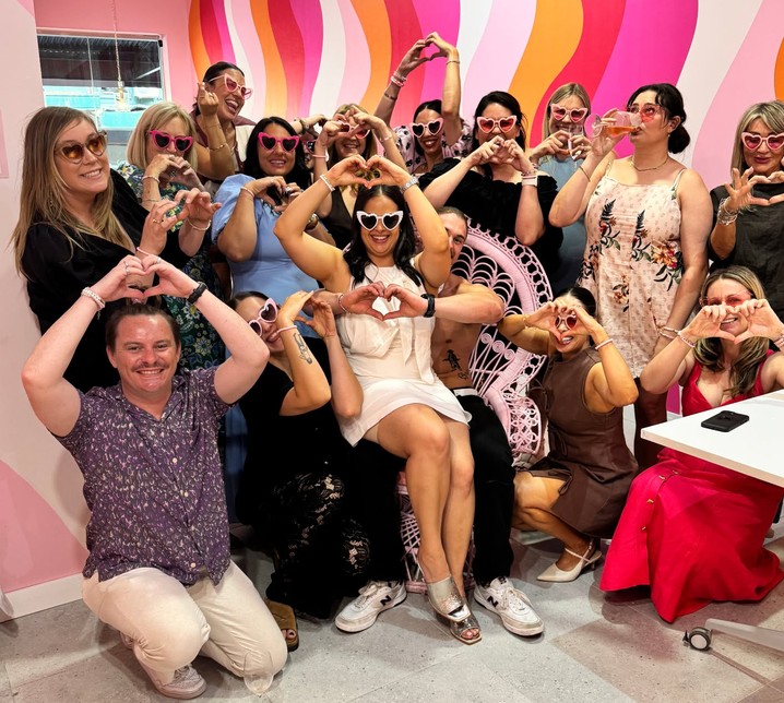 A hens crew in heart sunglasses making heart hands around the bride, with a shirtless man behind her chair