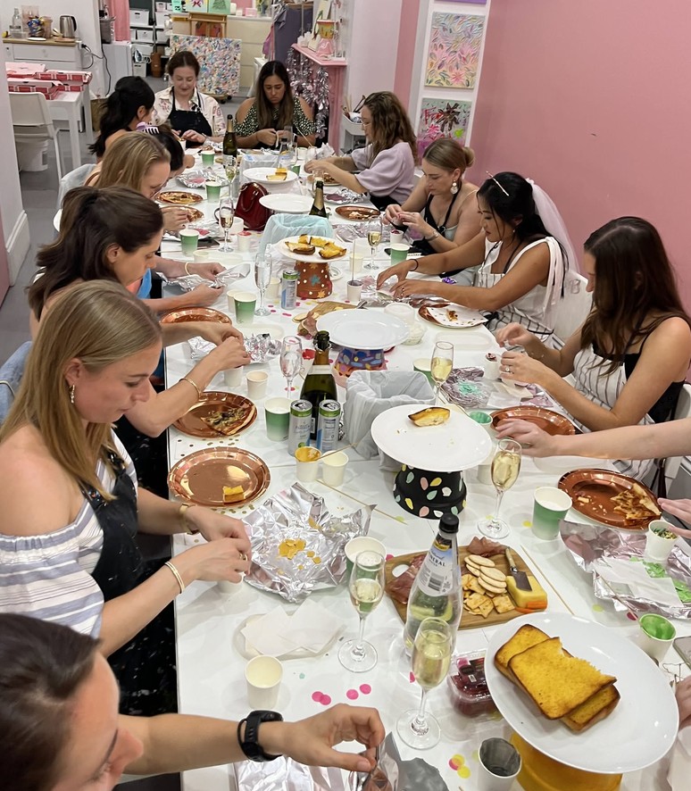 A long table of friends in aprons with plates of food, a grazing board, bubbly and the bride in a veil