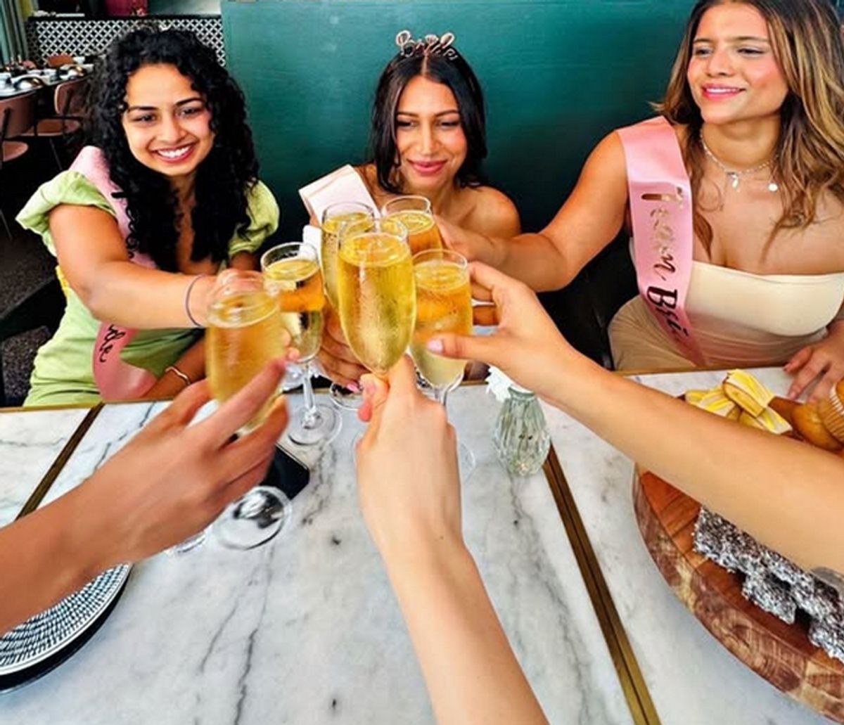 Hens in sashes and a tiara clinking glasses of bubbles across a marble table