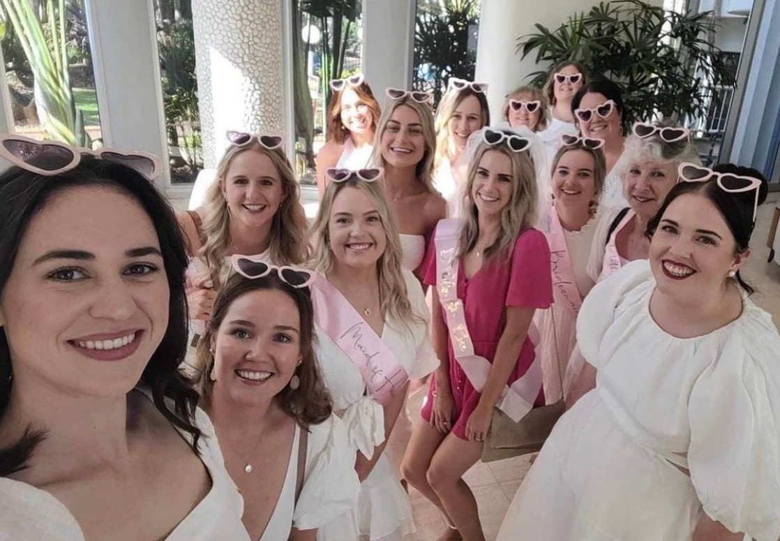 A big hens crew in white with pink heart sunglasses and sashes, the bride in pink in the middle, smiling for a group selfie