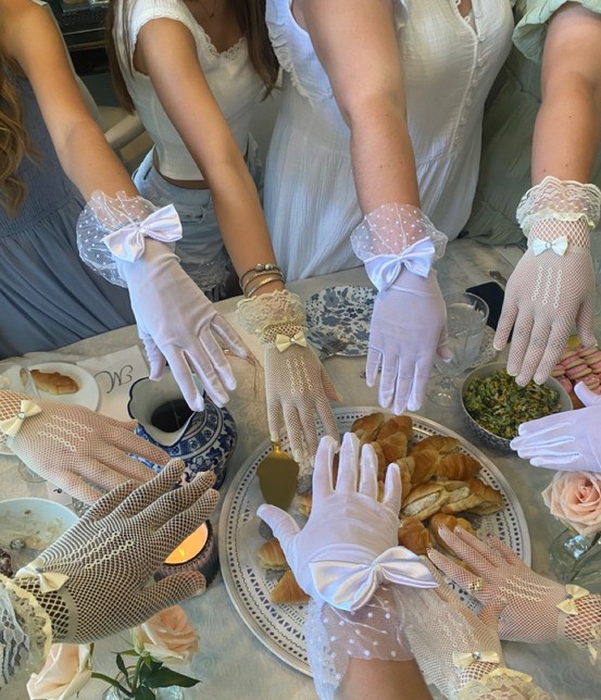 Hands in white lace and bow gloves meeting over a high tea table of pastries and roses