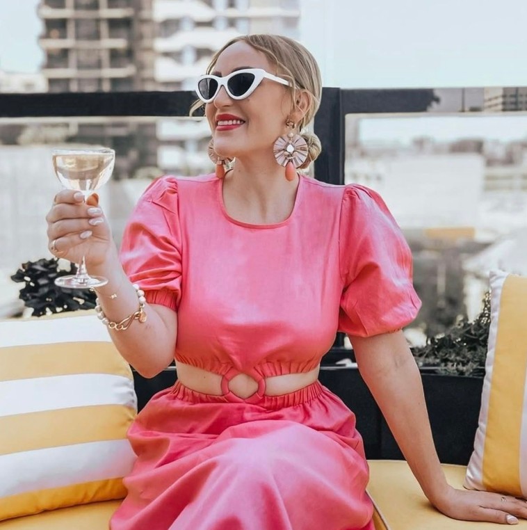 A woman in a pink dress and white cat-eye sunglasses raising a coupe on a rooftop